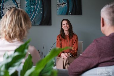 Vrouw in een oranje top zit lachend tegenover twee mensen in een interieur.