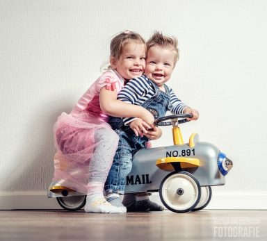 Twee kinderen, een meisje en een jongen, spelen op een speelgoed auto in een lichte kamer.