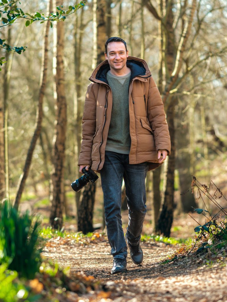 Man met camera wandelt glimlachend door een bosrijke omgeving, omgeven door bomen en groen.