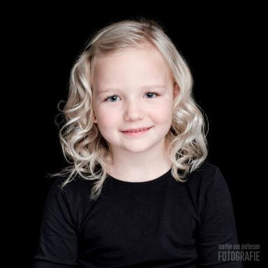 Blond meisje met krullend haar, lachend in een zwarte top op een donkere achtergrond.