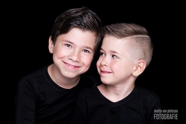 Twee jonge jongens met glimlachen, gekleed in zwart, tegen een zwarte achtergrond.