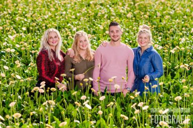 Groep van vier mensen, staand in een veld vol bloemen, met glimlachen en verschillende kledingstijlen.