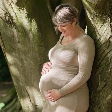 Zwangere vrouw in een beige jurk leunt tegen een boom en kijkt naar haar buik.