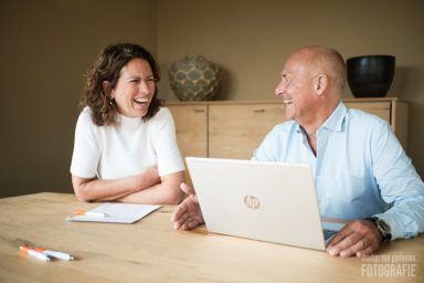 Twee personen lachend aan een tafel, met een laptop en notities voor hen.