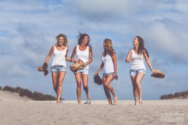 Vrouwelijke vrienden wandelen met blote voeten op het strand, lachend en met hoeden.