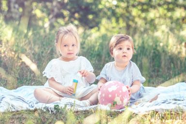 Twee jonge kinderen zitten op een deken in een groene, zonovergoten omgeving.