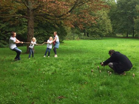 Vijf mensen in een park, met kinderen die aan een touw trekken terwijl een fotograaf fotografeert.