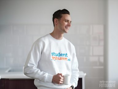 Jonge man in een witte trui met het logo "studententhuren.nl" en een mok in de hand.