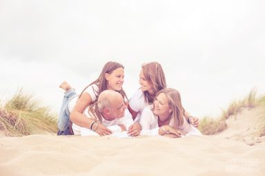 Een gezin met vier personen ligt lachend op een zandige bodem bij struiken.