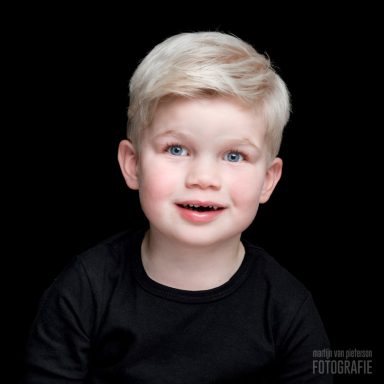 Blond jongetje met een lach, gekleed in zwart, tegen een zwarte achtergrond.