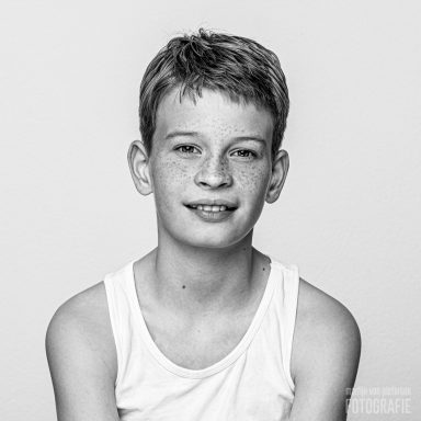 Een jongen met korte, lichte haren en een witte tanktop, glimlachend naar de camera.