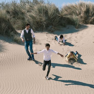 Een man en drie kinderen spelen op een zandstrand, met duinen op de achtergrond.