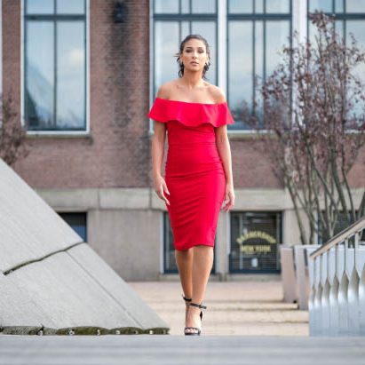 Vrouw in een rode off-shoulder jurk, lopend tussen moderne architectuur in Rotterdam.