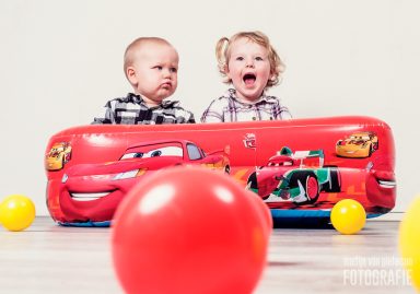 Twee kinderen in een opblaasbare auto met ballen eromheen, één lacht en de ander kijkt serieus.