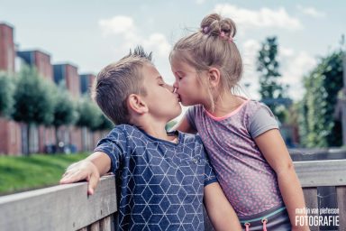 Twee kinderen kussen elkaar op een houten railing met een groene achtergrond.