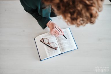Een persoon met gekruld haar leest en aantekeningen maakt in een boek met een bril erbij.