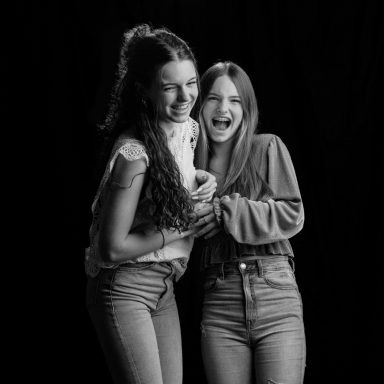 Twee lachende vrouwen met lange haren staan dichtbij elkaar voor een zwarte achtergrond.