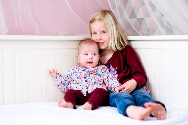 Meisje met lange blonde haren zit naast een baby in kleurrijke kleding op een bed.