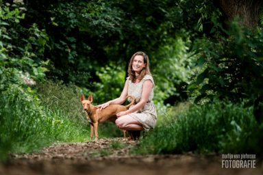 Een vrouw in een lichte jurk zit op een pad, naast een bruin hondje, omringd door groen.