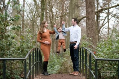 Een zwangere vrouw en een man houden een kind vast boven een brug in een bosrijke omgeving.