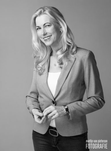 Blonde vrouw in een grijs jasje, glimlachend met een zelfverzekerde houding.